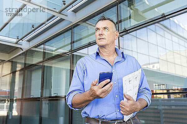 Nachdenklicher Geschäftsmann hält Smartphone und Dokument in der Hand  während er in der Stadt während einer Pandemie wegschaut