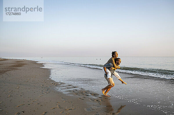 Fröhlicher Mann  der seine Freundin am Strand gegen den klaren Himmel bei Sonnenuntergang huckepack nimmt