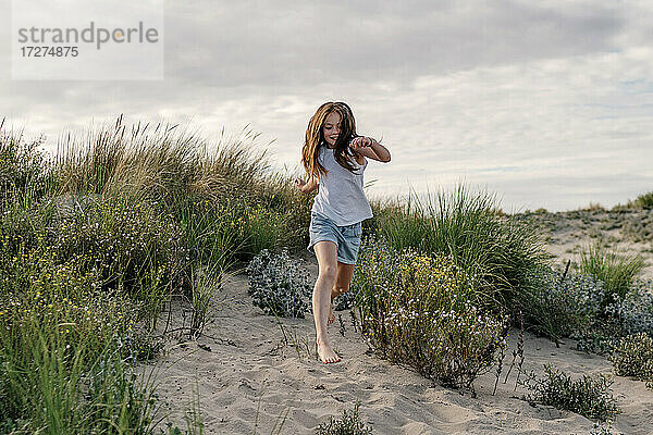 Fröhliches Mädchen läuft auf Sand am Strand gegen bewölkten Himmel