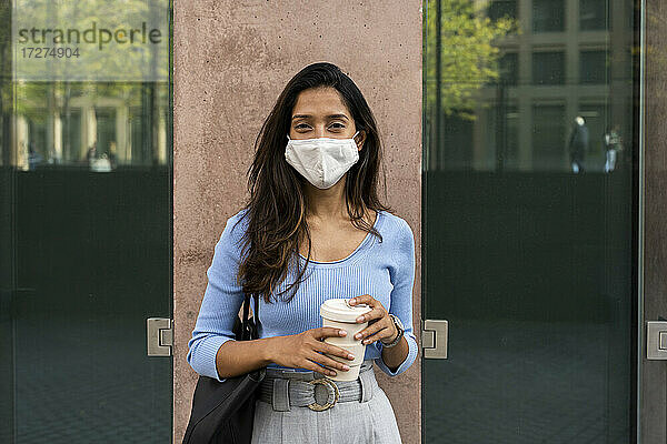 Junge Geschäftsfrau mit Schutzmaske  die einen Einweg-Kaffeebecher hält  während sie an einem Gebäude steht