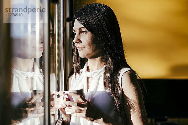 Frau schaut durch ein Fenster  während sie mit einer Kaffeetasse zu Hause steht