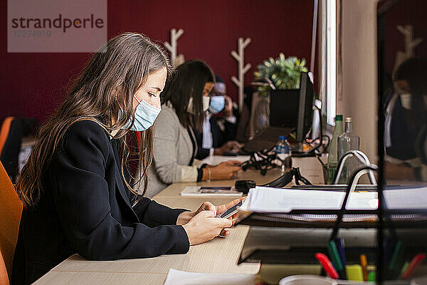 Junge Geschäftsfrau mit Gesichtsschutzmaske  die am Schreibtisch im Büro während einer Pandemie eine Textnachricht über ihr Smartphone schreibt