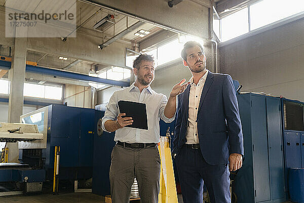 Ein Kollege benutzt ein digitales Tablet  während er mit einem Geschäftsmann in einer Fabrik diskutiert