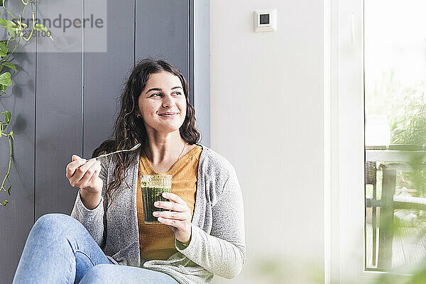 Lächelnde Frau mit Smoothie-Glas  die wegschaut  während sie zu Hause sitzt