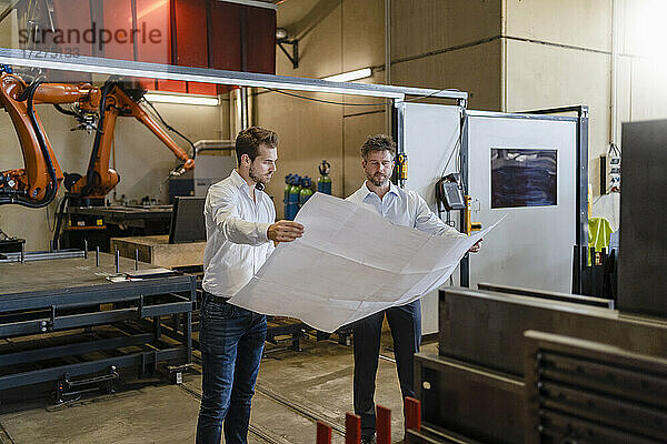 Geschäftsleute studieren den Bauplan eines Unternehmens  während sie vor einer automatisierten Maschine in einer Fabrik stehen