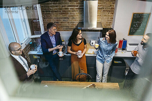 Hoher Blickwinkel von lächelnden Geschäftsleuten  die sich bei einem Kaffee in der Büroküche unterhalten  gesehen durch ein Glasfenster