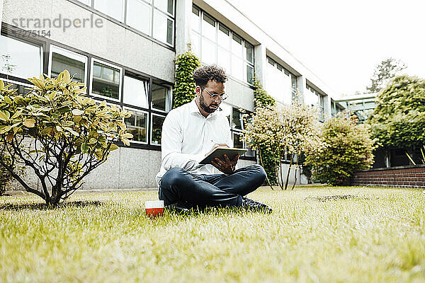 Geschäftsmann  der ein digitales Tablet benutzt  während er mit einer Kaffeetasse im Gras vor der Industrie sitzt
