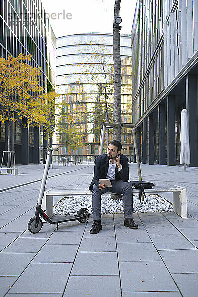 Unternehmer mit digitalem Tablet  der mit einem Elektroroller auf einer Bank in der Stadt sitzt