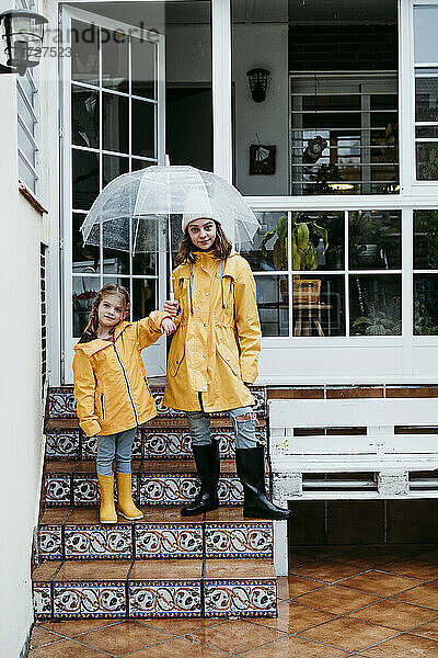 Schwestern mit Regenmantel und Regenschirm auf einer Treppe stehend