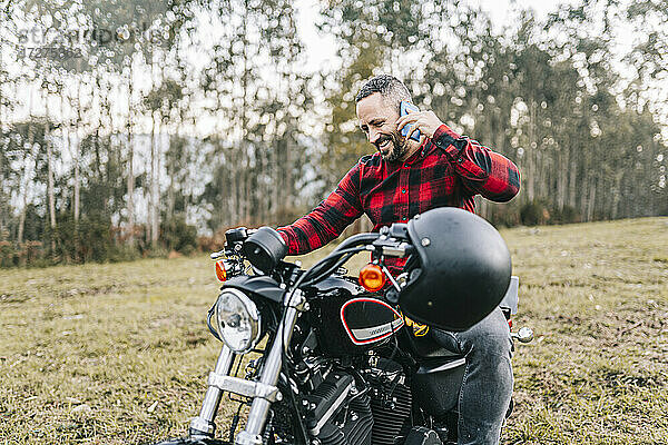 Glücklicher männlicher Biker  der auf einem Motorrad sitzend im Wald mit seinem Smartphone telefoniert