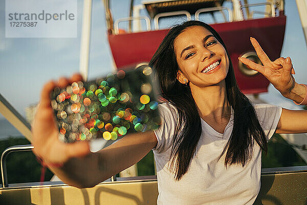 Schöne junge Frau  die ein Selfie mit ihrem Smartphone macht  während sie auf dem Riesenrad ein Friedenszeichen macht