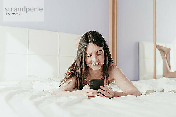 Lächelnde schöne Frau Text-Messaging  während auf dem Bett zu Hause liegen