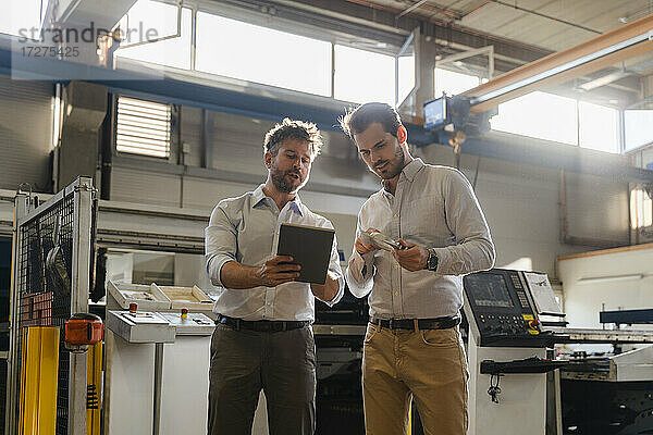 Geschäftsleute benutzen ein digitales Tablet  während sie in einer Fabrik ein Metallobjekt untersuchen