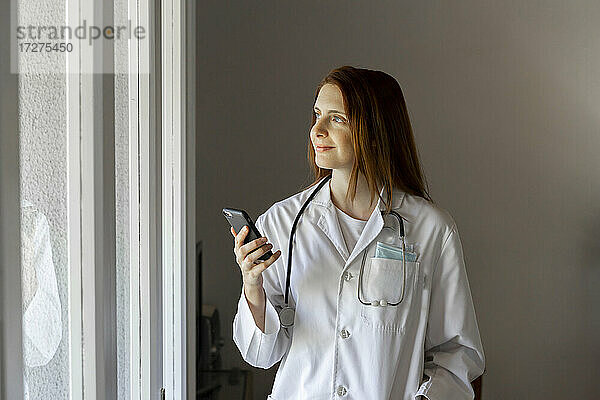 Lächelnde junge Ärztin schaut durch ein Fenster und hält ein Smartphone in der Hand