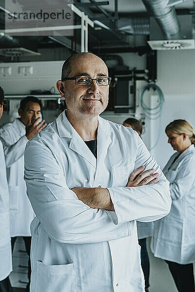 Lächelnder männlicher Wissenschaftler mit verschränkten Armen  der mit einem Mitarbeiter im Hintergrund in einem Labor steht