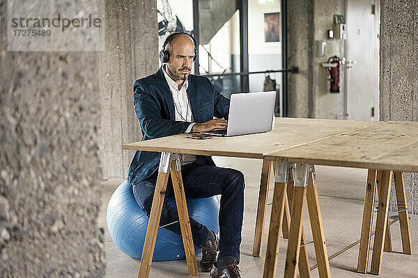 Geschäftsmann mit Kopfhörern  der einen Laptop benutzt  während er auf einem Fitnessball im Büro sitzt