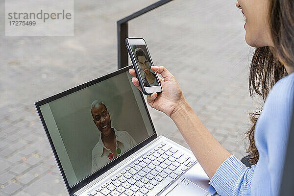 Geschäftsfrau führt während der COVID-19 im Freien sitzend Videogespräche über Laptop und Smartphone