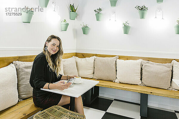 Lächelnde Frau  die ein Mobiltelefon benutzt  während sie am Tisch eines Cafés sitzt
