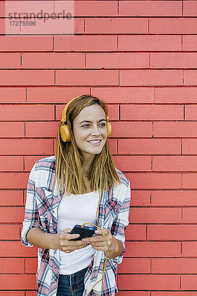 Junge Frau  die Musik über Kopfhörer hört  während sie ein Smartphone benutzt  das an einer Backsteinmauer steht