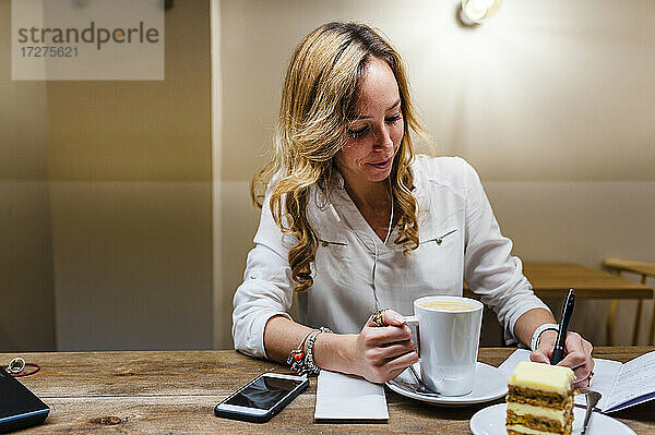Unternehmerin schreibt in ein Notizbuch  während sie am Tisch Kaffee trinkt
