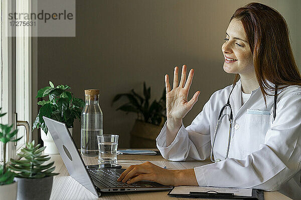 Junge Ärztin winkt dem Patienten zu  während sie eine Online-Konsultation von zu Hause aus durchführt