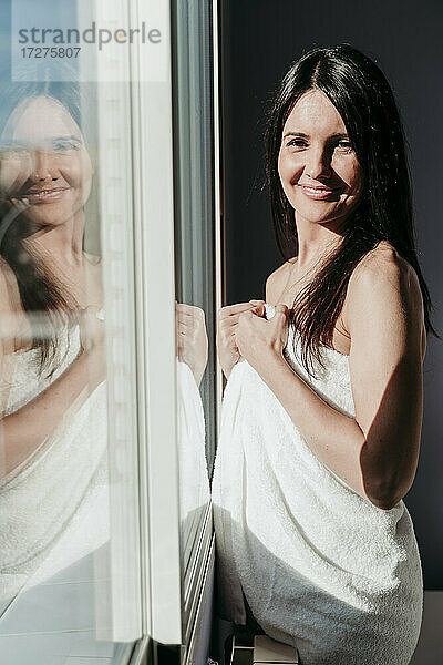 Lächelnde schöne Frau  eingewickelt in ein Handtuch  steht am Fenster zu Hause