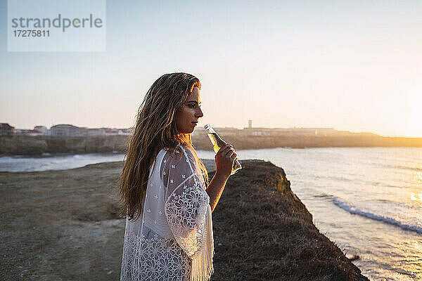 Junge Frau mit kleiner Weißweinflasche  die am Strand steht und wegschaut