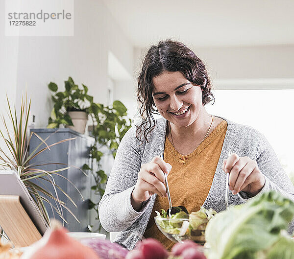 Lächelnde Frau isst Salat  während sie zu Hause sitzt