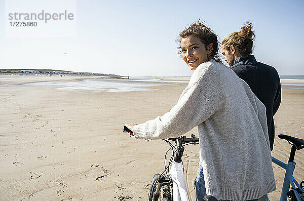 Lächelnde schöne Frau  die mit ihrem Freund auf dem Fahrrad spazieren geht  während sie über die Schulter auf den Strand gegen den klaren Himmel an einem sonnigen Tag schaut