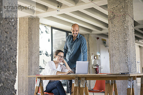 Lächelnde Frau  die einen Laptop benutzt  während ein Mann am Tisch im Büro steht