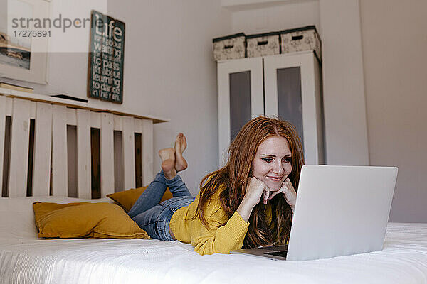 Lächelnde Frau auf der Vorderseite liegend mit der Hand am Kinn  die zu Hause einen Laptop benutzt
