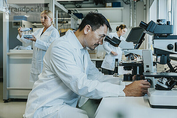 Männlicher Wissenschaftler sitzt am Mikroskop  während ein Mitarbeiter im Hintergrund im Labor arbeitet