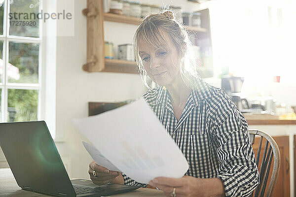 Ältere Frau mit Papier in der Hand sitzt mit Laptop in der Küche zu Hause