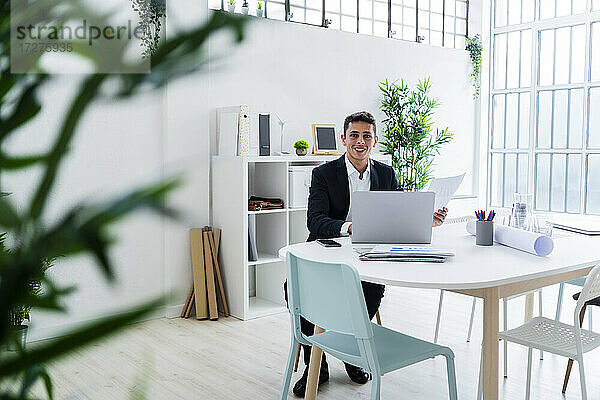 Lächelnde männliche Fachkraft sitzt mit Laptop am Schreibtisch in einem kreativen Büro