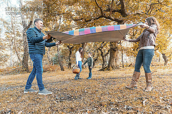 Eltern legen eine Picknickdecke aus  während die Kinder im Herbst im Park stehen