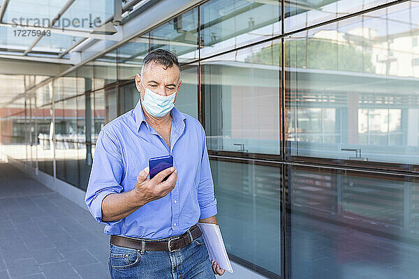 Männlicher Unternehmer benutzt sein Smartphone während eines Spaziergangs mit einem Dokument in der Stadt während einer Pandemie