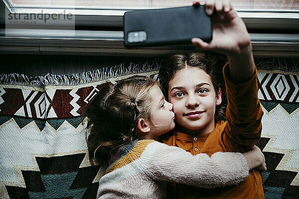 Mädchen küsst Schwester  während sie ein Selfie mit dem Handy zu Hause macht