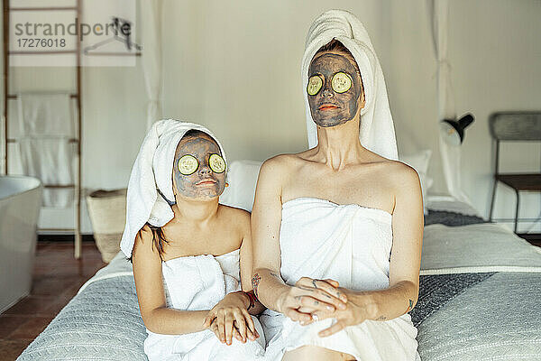 Mutter und Tochter im Handtuch mit Gesichtsmaske und Gurkenscheibe auf den Augen sitzen zu Hause