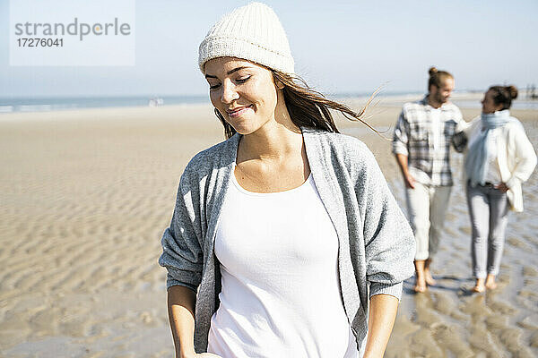 Junge Frau lächelt beim Spaziergang mit Freund und Mutter im Hintergrund am Strand
