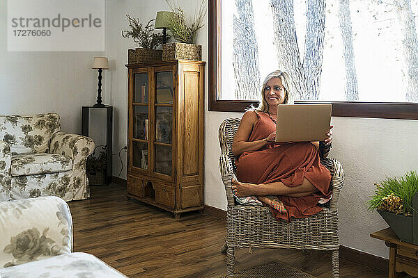 Lächelnde blonde Frau  die wegschaut  während sie mit einem Laptop auf einem Korbsessel sitzt