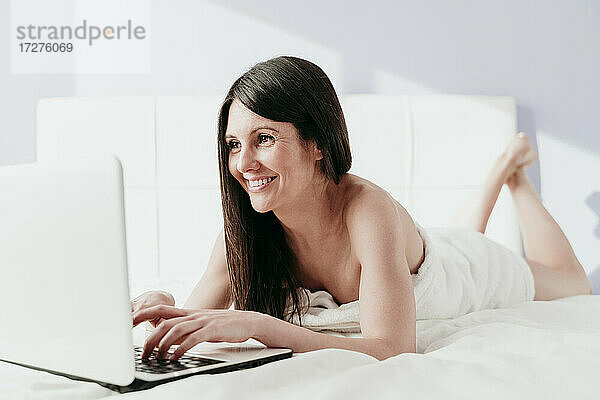 Lächelnde schöne Frau  die einen Laptop benutzt  während sie zu Hause auf dem Bett liegt