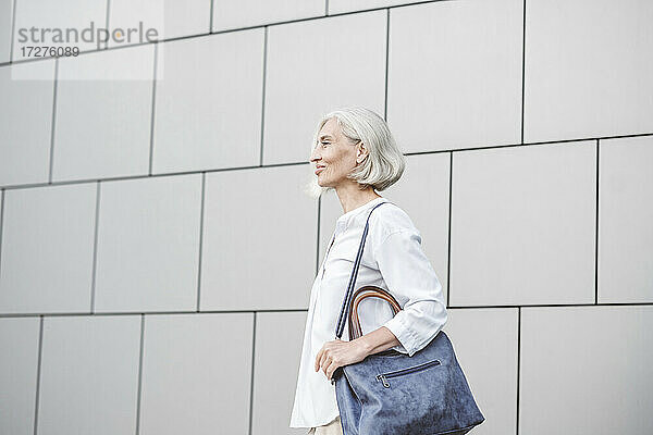 Lächelnde Geschäftsfrau mit Handtasche schaut weg  während sie an der Wand steht