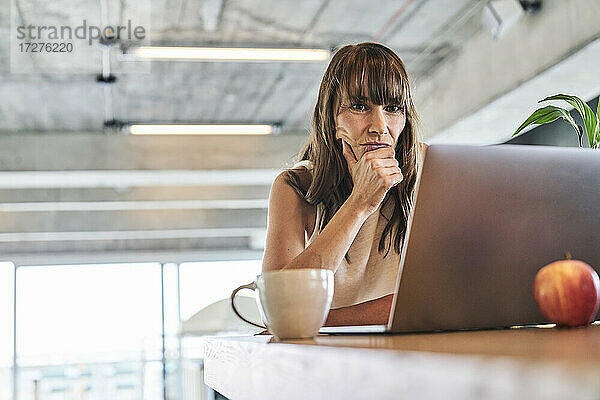 Nachdenkliche Frau mit Hand am Kinn  die einen Laptop benutzt  während sie zu Hause sitzt