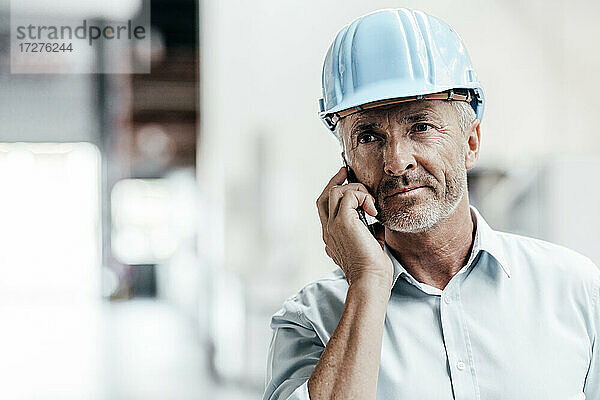 Reifer männlicher Ingenieur mit Schutzhelm  der wegschaut  während er in einer Fabrik mit einem Mobiltelefon spricht