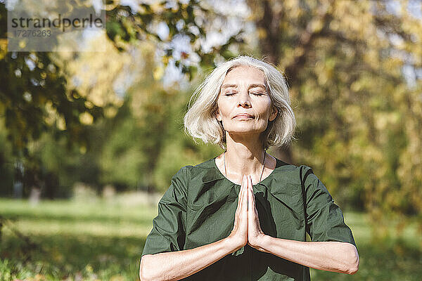 Reife Frau sitzt mit verschränkten Händen beim Yoga im Park