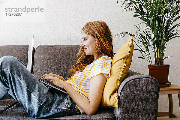 Rothaarige Frau auf dem Sofa liegend mit Laptop zu Hause