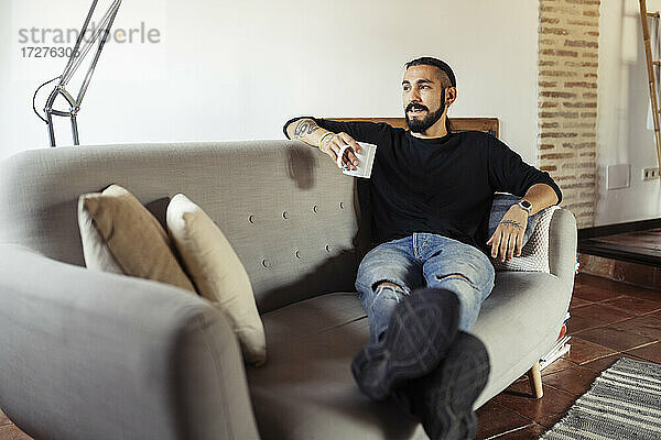 Mann hält Kaffeetasse  während er zu Hause auf dem Sofa sitzt