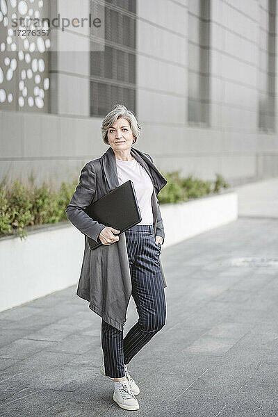 Selbstbewusste Geschäftsfrau  die einen Laptop hält und mit der Hand in der Tasche vor einem Gebäude steht