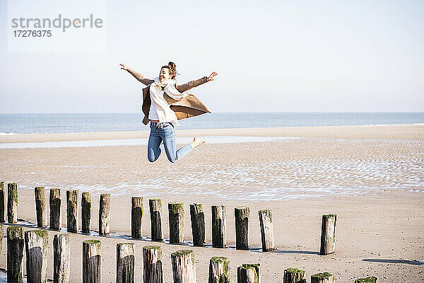 Glückliche junge Frau mit ausgestreckten Armen  die an einem sonnigen Tag von einem Holzpfosten am Strand springt