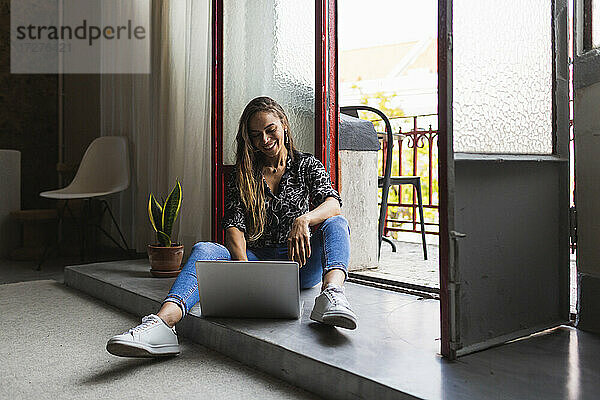 Lächelnde kreative Geschäftsfrau  die am Laptop arbeitet  während sie an der Tür im Büro sitzt